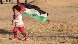 A Palestinian girls runs with the national flag, as protesters burn tires at the site of a tent protest on April 8, 2018, on the Israel-Gaza border east of Rafah in the southern Gaza Strip
