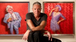 Bill Leak poses in front of two painting of Australian comedian Barry Humphries