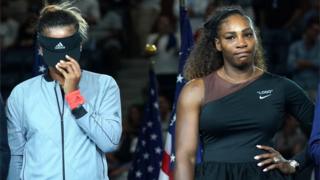 Naomi Osaka pulls her visor down covering her face, Serena Williams stands hand on one hip