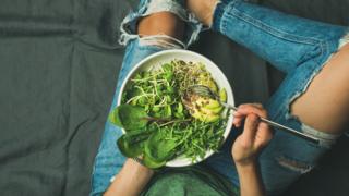 Young woman sitting holding a bowl full of spinach, rocket and avocado.