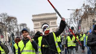 A man wearing an Anonymous mask and a yellow vest and holding a Star Wars Jedi light laser takes part in a demonstration to protest against rising costs of living they blame on high taxes, by the Arc de Triomphe in Paris, on December 15, 2018